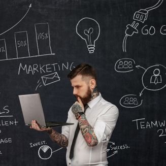 Tattooed bearded techer with laptop over school desk with formulas.